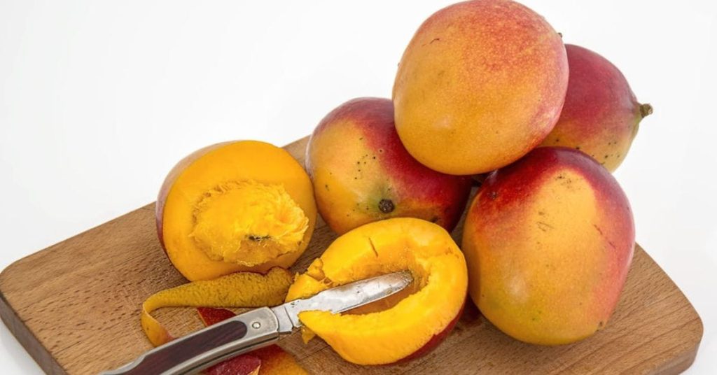 Manga cortada sobre tábua de madeira com faca e frutas inteiras ao fundo
