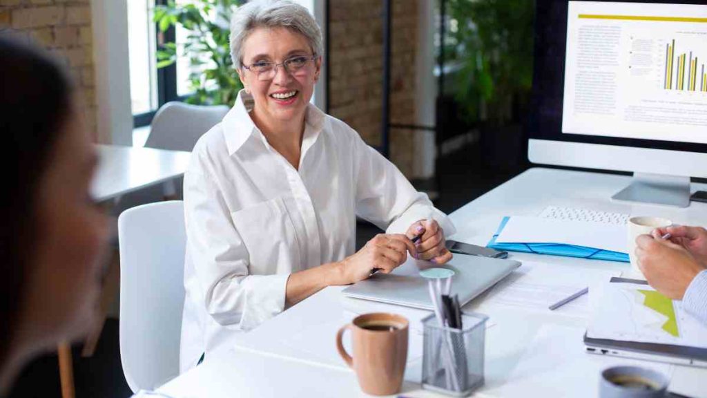 Mulher sorrindo segurando uma caneta, com um notebook na mesa em companha com pessoas em segundo plano, representando a empregabilidade de mulheres 50+