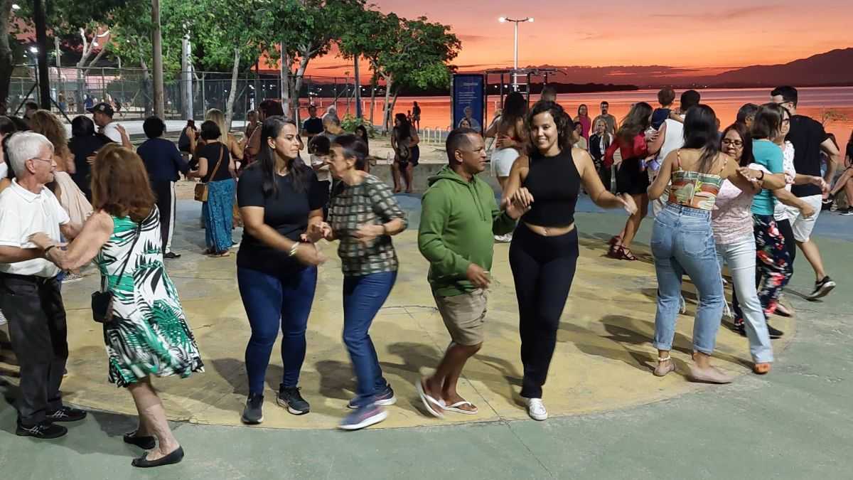 Grupo de pessoas de todas as idades dançando em praça pública ao pôr do sol na Ilha do Governador, durante o projeto Dançando na Praça com Leandro Azevedo.