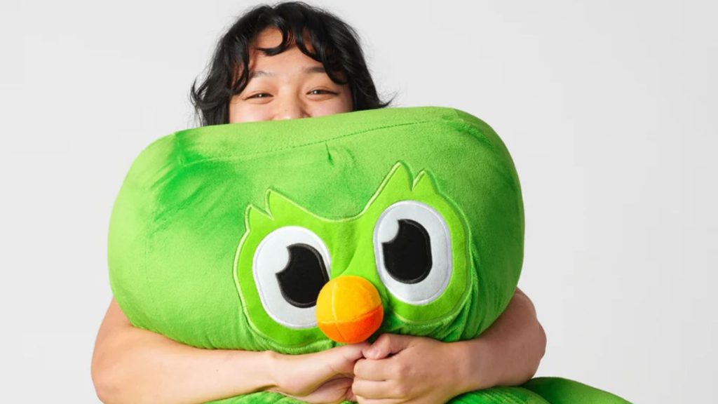 Jovem sorridente abraça uma pelúcia gigante do Duo, a icônica coruja verde do Duolingo, representando o carinho dos usuários e a febre do Duolingo no mundo.