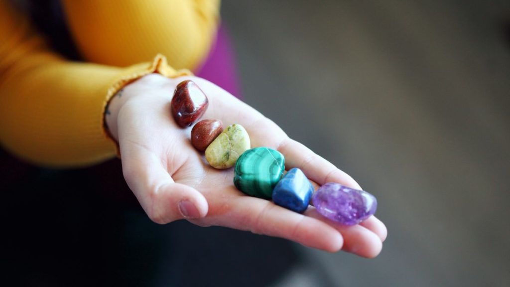 Mão segurando sete pedras coloridas representando o poder dos 7 chakras, simbolizando equilíbrio energético e cura espiritual.