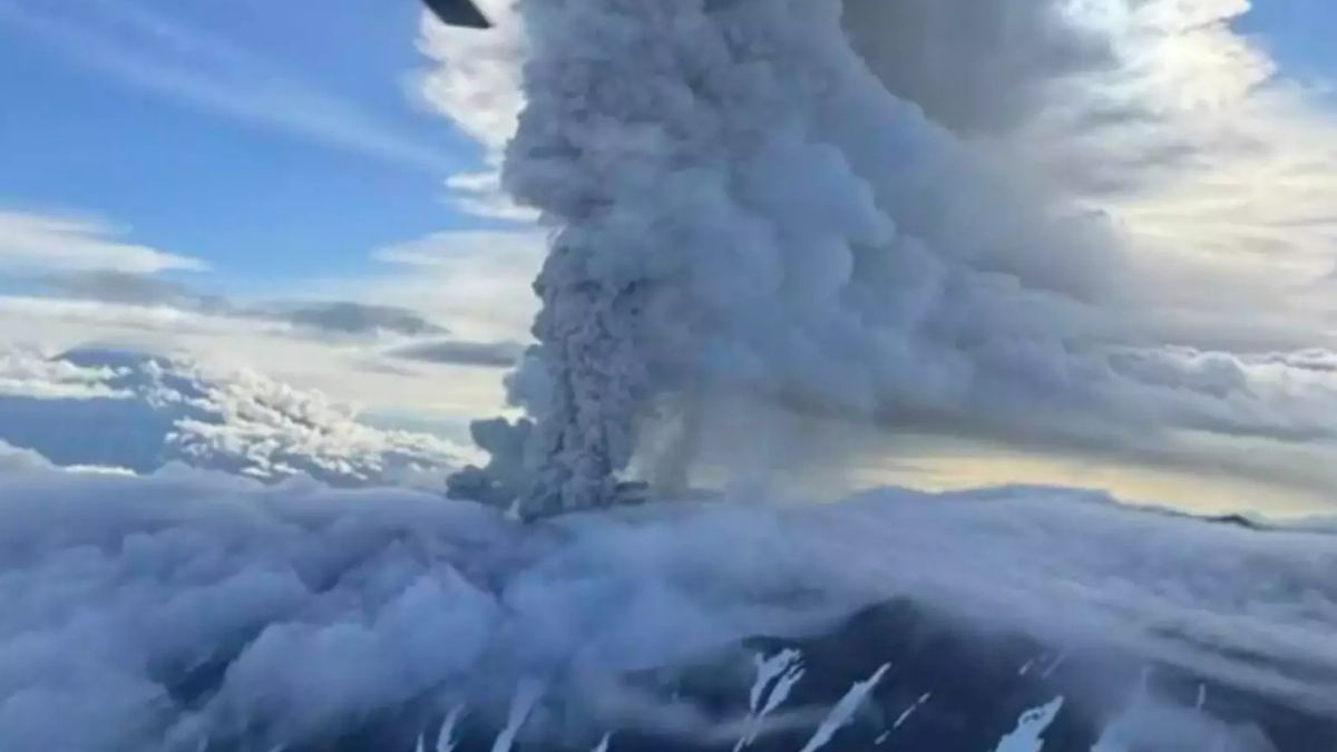 Imagem aérea do vulcão Krasheninnikov em erupção, com coluna de cinzas atingindo o céu e fluxo de lava visível.