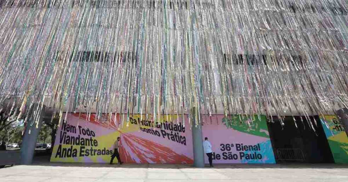 Fachada da Bienal de São Paulo com instalação artística de fitas metálicas coloridas suspensas, criando um efeito de cortina cintilante. Ao fundo, o painel da entrada exibe o título da edição “Nem todo viandante anda estradas – Da humanidade como prática”.