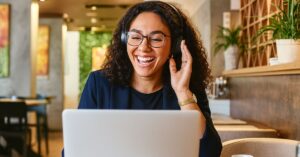 Mulher sorridente com fones de ouvido trabalhando em laptop em um café iluminado, simbolizando equilíbrio emocional no Dia Mundial da Saúde Mental.