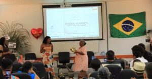 Idosa apresenta o TCC na UFNT segurando saxofone ao lado da orientadora, durante defesa de Maria de Fátima TCC