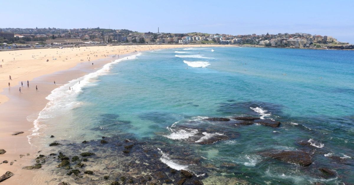 Praia de Bondi em Sydney, com faixa de areia clara, mar azul e área urbana ao fundo