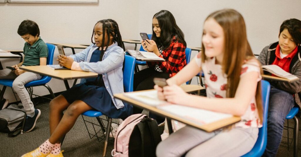 Crianças usando celular em sala de aula, ilustrando debates sobre segurança para crianças nas redes.
