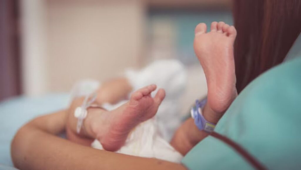 Recém-nascido em UTI neonatal sendo monitorado, com foco nos pés do bebê durante cuidados médicos