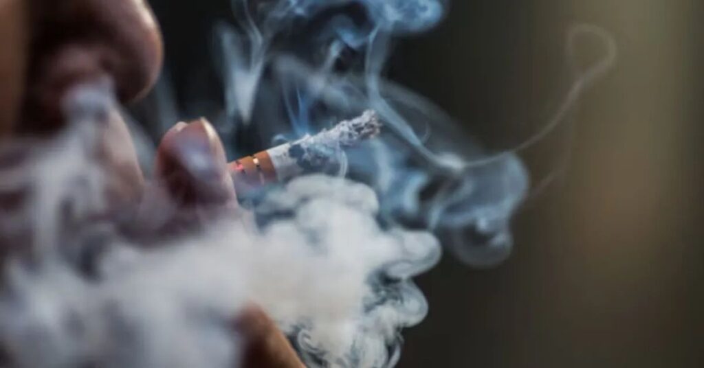 Pessoa fumando cigarro enquanto fumaça se espalha, imagem que ilustra os benefícios de parar de fumar e os riscos do tabagismo para a saúde.