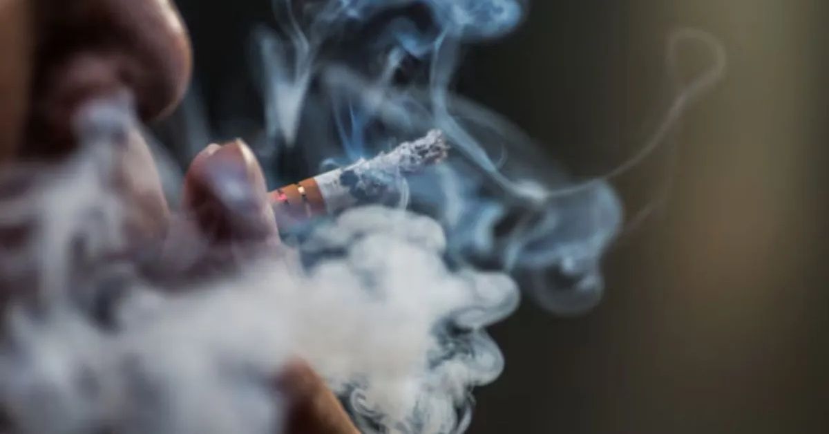 Pessoa fumando cigarro enquanto fumaça se espalha, imagem que ilustra os benefícios de parar de fumar e os riscos do tabagismo para a saúde.