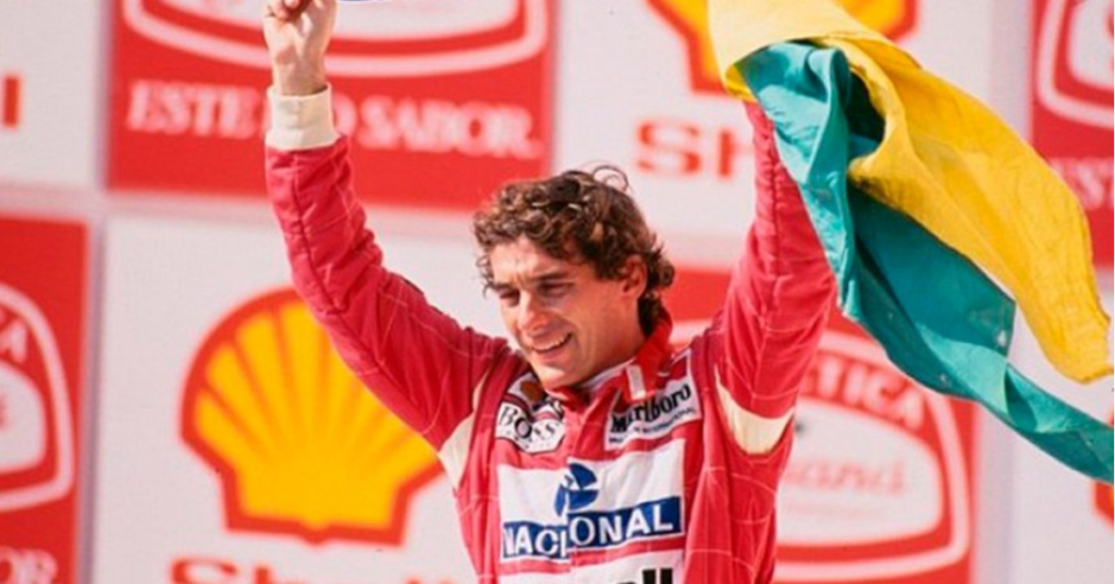 Ayrton Senna ergue a bandeira do Brasil no pódio após uma vitória na Fórmula 1, usando macacão vermelho de corrida.