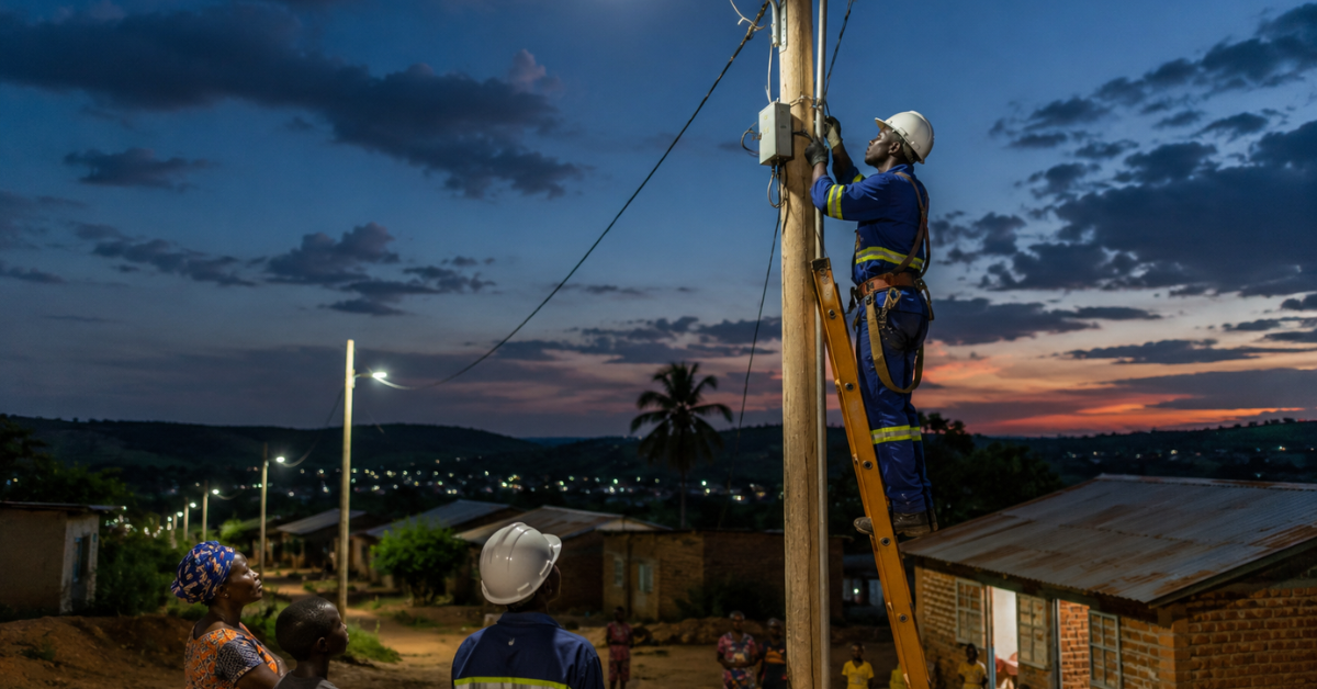 Técnico realiza instalação em poste de energia enquanto moradores acompanham a expansão da eletrificação em uma comunidade africana.