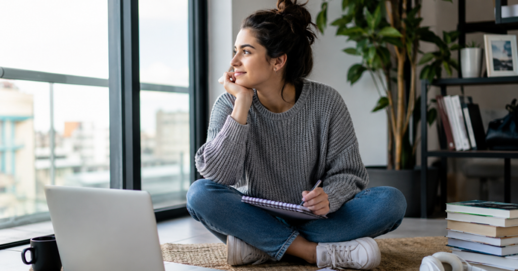 Jovem sentada no chão de casa com notebook, caderno e livros ao redor, em um ambiente de estudo e planejamento pessoal.