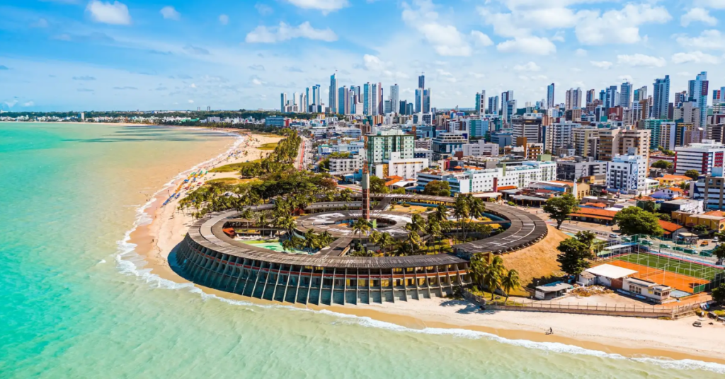 Vista aérea de João Pessoa, com praia, mar e prédios da orla da capital paraibana.
